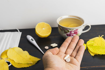 Male hand holds medicine pill and capsule above table with tea cup with lemon, thermometer, medical mask and yellow leaves. Concept of treatment autumn cold, flu and coronavirus.の写真素材