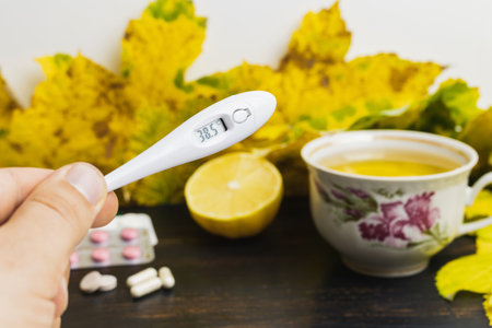Male hand holds digital thermometer indicates high temperature 38.5, above table with tea cup, lemon, capsules, pills and yellow leaves. Concept of treatment fever, autumn cold, flu and coronavirus.の写真素材