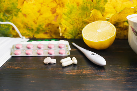 Capsules, pills, medicine in a blister pack, thermometer, lemon on a wooden table with yellow leaves. Concept of treatment autumn cold, flu and coronavirus.の写真素材