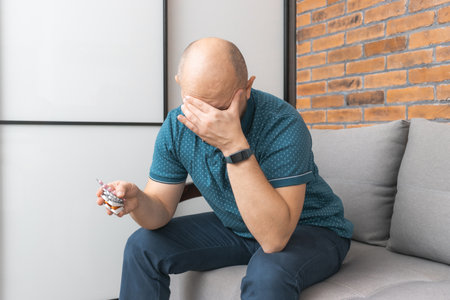 Sad and depressed man sitting on a sofa, he has a headache or migraine, holding in a hand various blister packs with pills and medicines.の写真素材
