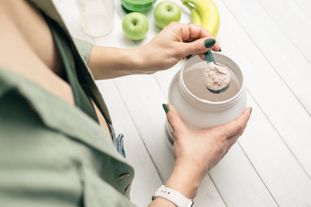 Woman in jeans and shirt holding measuring spoon with portion whey protein powder above plastic jar on white wooden table with shaker, banana and apple fruit. Process of making protein drink.の写真素材