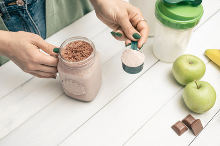Young woman in jeans and shirt holding measuring spoon with protein powder, glass jar of protein drink cocktail, milkshake or smoothie above white wooden table with chocolate pieces, bananas, apples.の写真素材