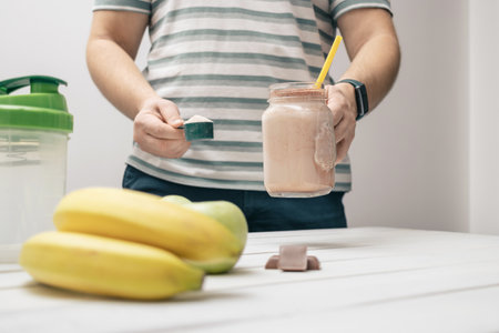 Man holding measuring spoon with protein powder, glass jar of protein drink cocktail, milkshake or smoothie above white wooden table with chocolate pieces, bananas, apples.の写真素材