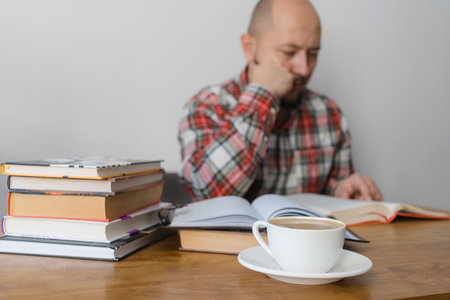 Man reading a book, studying or working, sitting at the table with stack of books and cup of coffee, focus on foreground.の写真素材