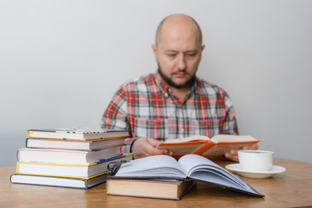 Man reading a book, studying or working, sitting at the table with stack of books and cup of coffee, focus on foreground.の写真素材