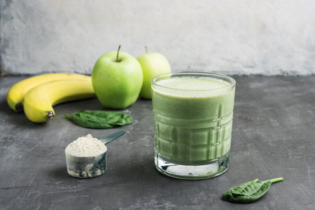 Green detox smoothie in a glass, measuring spoon with protein powder, blended vegetarian drink from spinach leaves, apple and banana fruit, healthy eating concept.の写真素材