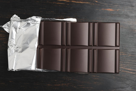 Delicious dark chocolate bar wrapped in foil on a dark wooden background, top view.の写真素材