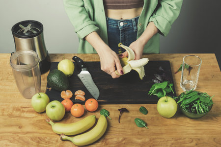 Woman peeling banana, preparing healthy vegan smoothie with spinach leaves, apple, tangerine and avocado on a table.の写真素材