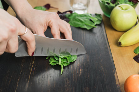 Woman cutting green spinach leaves, preparing healthy vegan smoothie with spinach, banana, apple and avocado in a blender on a table.の写真素材