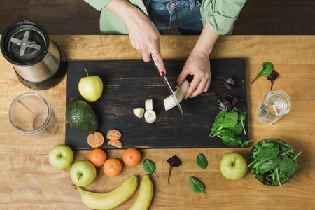 Woman cutting banana, preparing healthy vegan smoothie with spinach leaves, apple and avocado on a...