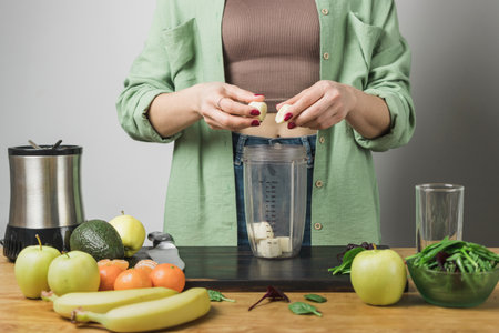 Woman holding banana pieces, preparing healthy vegan smoothie with spinach, apple and avocado in a blender on a table.の写真素材