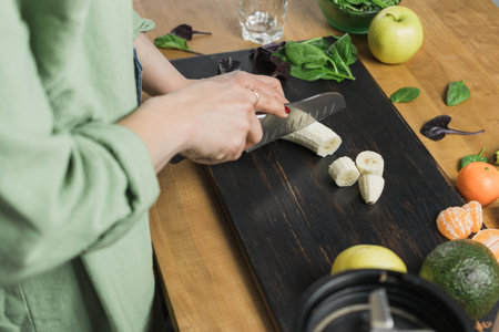Woman cutting banana, preparing healthy vegan smoothie with spinach leaves, apple and avocado on a table.の写真素材