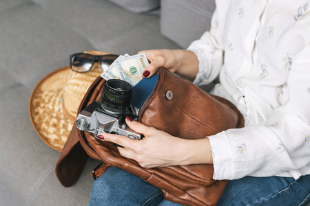 Woman packing leather backpack with retro camera, passport and dollars cash currency, straw hat,...