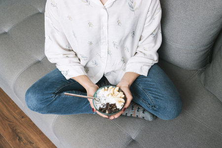 Woman sitting on a sofa and eating healthy food for breakfast, cereal, granola, muesli, oatmeal with banana, dark chocolate and yogurt in a bowl.の写真素材