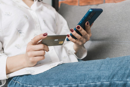 Unrecognizable woman sitting on couch, holding credit card and using mobile phone, smartphone for...
