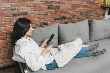 Beautiful young woman covered with blanket, lying on a couch, holding credit card and using mobile...
