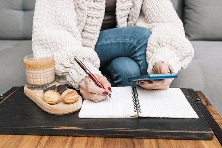 Unrecognizable woman sitting on a couch at home, using mobile phone or smartphone and writing in notepad on the table with glass of coffee drink and dessert.の写真素材