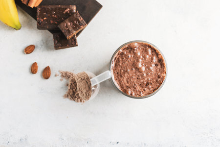 Glass with chocolate protein drink, milkshake smoothie on white table with bananas, protein powder in measuring spoon, protein bar, almond nuts, top view.の写真素材