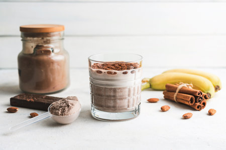 Glass with chocolate protein drink, milkshake smoothie on white table with bananas, protein powder in measuring spoon, protein bar, almond nuts and cinnamon sticks.の写真素材