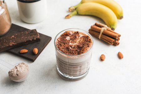 Glass with chocolate protein drink, milkshake smoothie on white table with bananas, protein powder in measuring spoon, protein bar, almond nuts and cinnamon sticks.の写真素材