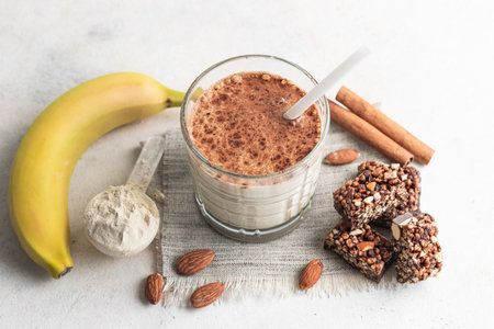 Glass with protein drink, healthy milkshake smoothie on white table with bananas, protein powder in measuring spoon, protein bar, almond nuts and cinnamon sticks.の写真素材