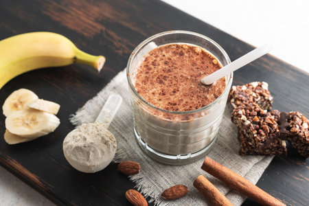Glass with protein drink, healthy milkshake smoothie on wooden board with bananas, protein powder in measuring spoon, protein bar, almond nuts and cinnamon sticks.の写真素材