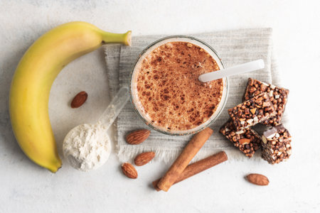 Glass with protein drink, healthy milkshake smoothie on white table with bananas, protein powder in measuring spoon, protein bar, almond nuts and cinnamon sticks, top view.の写真素材