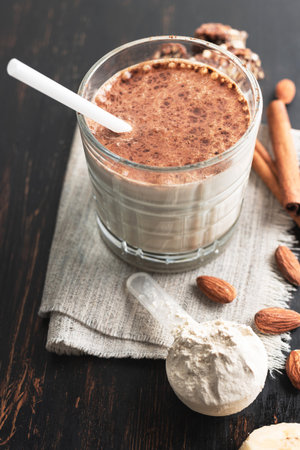 Glass with protein drink, healthy milkshake smoothie on wooden board with protein powder in measuring spoon, almond nuts and cinnamon sticks.の写真素材