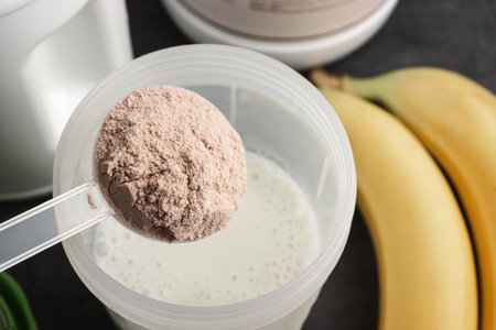 Plastic measuring spoon puts whey protein powder into a shaker with milk on a dark background with...