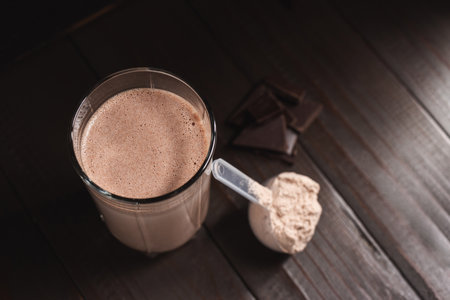 Plastic measuring spoon with whey protein powder, milkshake cocktail in a glass, blended protein drink and chocolate cubes on a dark wooden background.の写真素材