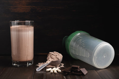Protein milkshake cocktail in a glass, plastic measuring spoon with whey protein powder, white pills or capsules of amino acids, chocolate cubes on a dark wooden background.の写真素材