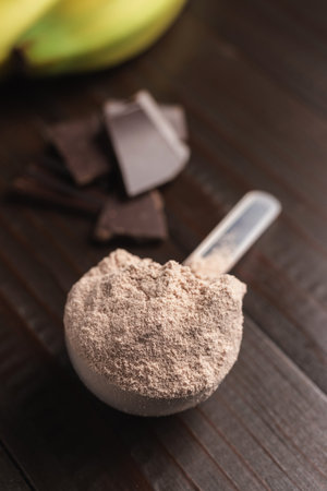 Plastic measuring spoon with whey protein powder, chocolate cubes and banana fruit on a dark wooden background.の写真素材