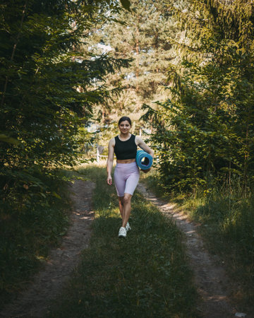 A young sporty woman with fitness mat walking in the summer park, morning workout concept.