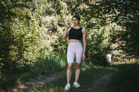 A young sporty woman drinking water from a bottle in the summer park, morning workout and hydration...