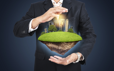 Businessman in suit keeping green island with skycraper city in hands. Care city concept.の写真素材