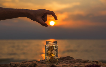 Saving money concept, Male hand putting sun (money coin) stack growing business with selective focus, shallow depth of field and sunset background.の写真素材