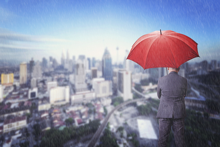 Businessman with umbrella standing over rain and blur cityscape background. Business, protection, crisis concept.の写真素材