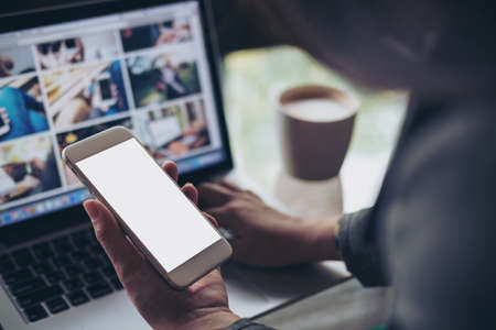 Mockup image of a business woman holding mobile phone with blank white screen while using laptop with coffee cup on the table in officeの写真素材