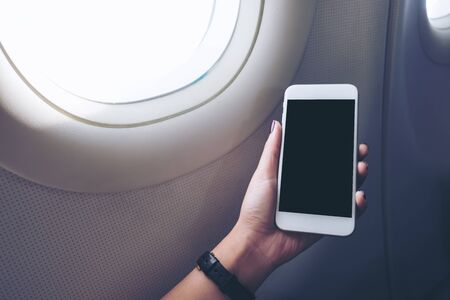 Mockup image of a hand holding and looking at white smart phone with black screen next to an airplane window with clouds and sky backgroundの写真素材