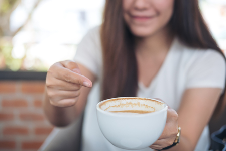 Closeup image of Asian woman holding and pointing at hot coffee with smiley face in coffee shopの写真素材