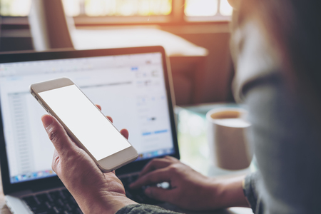 Mockup image of a woman holding mobile phone with blank white screen while using laptop with coffee cup on the table in officeの写真素材