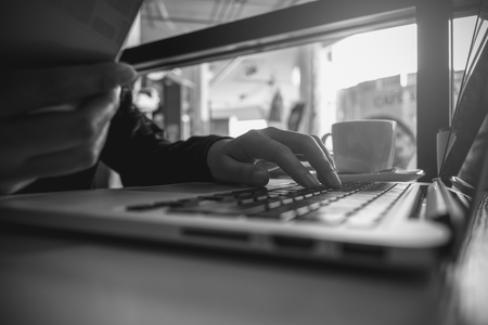 Black and white image of a business woman's hands holding mobile phone and typing on laptop keyboard on wooden tableの写真素材
