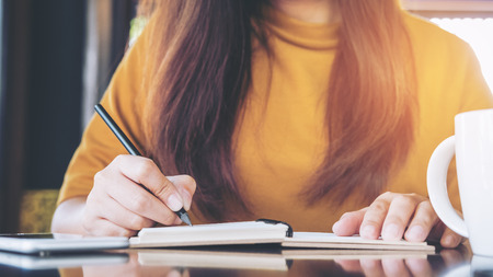 Close up image of a woman writing on notebook with coffee mug on wooden table in cafe