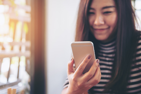A beautiful Asian girl with smiley face holding and using smart phone in modern cafeの写真素材