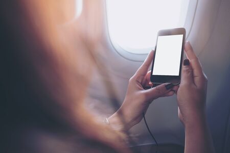 Mockup image of a woman holding and looking at black smart phone with black white screen next to an airplane window with clouds and sky backgroundの写真素材