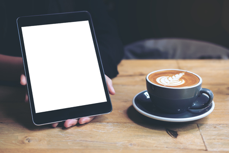 Mock up image of a woman's hands showing black tablet with white blank screen and coffee cup on wooden table in cafeの写真素材