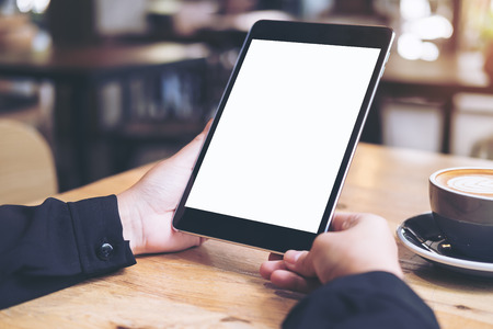 Mock up image of business woman's hands holding black tablet with white blank screen and coffee cup on wooden table in cafeの写真素材