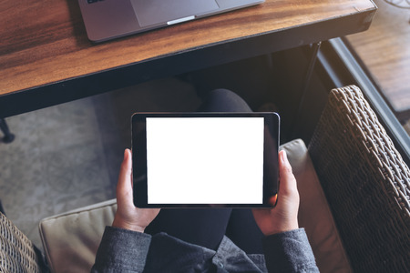 Mockup image of woman's hands holding black tablet pc with blank white screen on thigh with laptop...