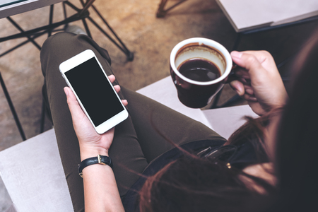 Mockup image of a woman holding white mobile phone with blank black screen on thigh and drinking coffee in modern loft cafeの写真素材