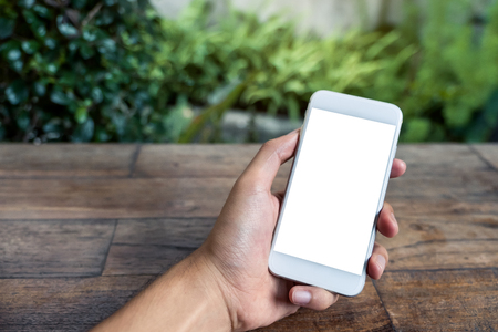 Mockup image of a man's hand holding and using white smart phone with blank screen on wooden table and green nature backgroundの写真素材